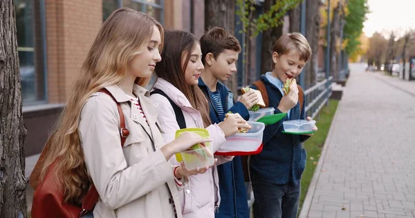 Retrato de adolescentes de escuela caucásica felices de pie al aire libre con recipientes de comida y sonriendo mientras come sándwiches. Alegre niños y niñas alumnos con mochilas tomando bocadillos cerca de la escuela en el descanso — Foto de Stock