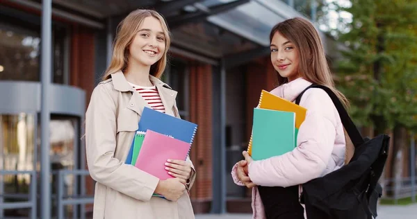 Portret van vrolijke mooie jonge meisjes scholieren met copybooks die in goed humeur op straat staan en glimlachen naar de camera. Blanke gelukkige tienervriendinnen. Concept klasgenoten — Stockfoto