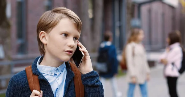 Primer plano retrato de niño caucásico feliz escuela de pie al aire libre y hablando en el teléfono inteligente. Alegre estudiante masculino con mochila llamando al celular en la calle. Concepto de alumnos —  Fotos de Stock