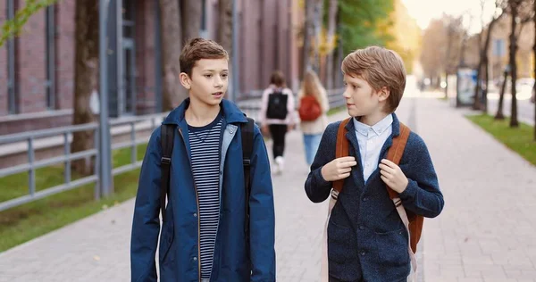 Caucásico feliz agradable macho escuela estudiantes caminando en la calle después de la escuela y charlando. Retrato de niños alegres alumnos con mochilas de buen humor hablando al aire libre en la ciudad. Concepto de compañeros —  Fotos de Stock