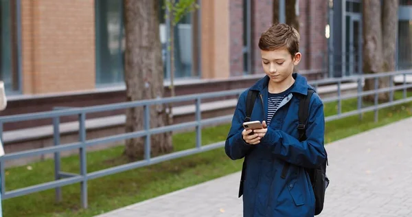 Portrait d'un écolier heureux marchant dans la rue jusqu'à l'école et tapant sur son téléphone portable. Elève homme agitant la main à des camarades de classe filles à l'extérieur. Textos étudiants et tapotements sur smartphone. Concept de loisirs — Photo