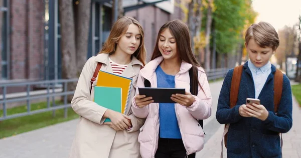 Retrato de adolescentes caucásicos caminando y buscando en internet en diferentes dispositivos. Chica feliz tocando en la tableta al aire libre. Chico mensajes de texto en el teléfono inteligente en la calle en la ciudad mientras va a la escuela — Foto de Stock