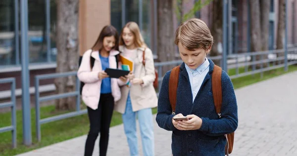 Portré kaukázusi iskolás fiúról, aki iskolába megy és mobilon gépel a szabadban. Férfi tanuló sms-ezik és kopog az okostelefonon. Lányok tapogatóznak a háttérben. A pupilla fogalma — Stock Fotó