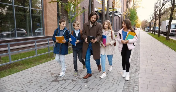 Caucasian children with copybooks walking to school with male adult teacher on street in city. Happy man educator with girls and boys students tapping on tablet outdoors. Education concept — Stock Photo, Image