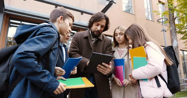 Retrato de niños caucásicos con coloridos libros de texto de pie alrededor del educador hombre cerca de la escuela en la calle de la ciudad. Feliz profesor masculino escribiendo en la tableta y mostrando algo a las niñas y niños alumnos — Foto de Stock