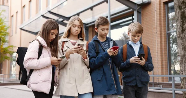 Blanke geconcentreerde tieners die buiten staan en typen op mobieltjes. Mooi meisje leerling browsen op smartphone in de buurt van school. Jongensscholier sms 't op mobiele telefoon. Vrienden concept — Stockfoto