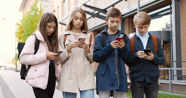 Les adolescents caucasiens concentrés debout à l'extérieur et tapant sur les téléphones cellulaires. Belle élève fille naviguant sur smartphone près de l'école. Jeune garçon écolier textos sur téléphone portable. Concept d'amis — Photo