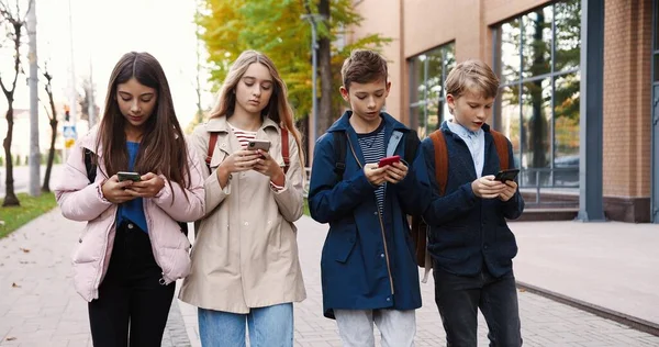 Beyaz tenli gençler dışarı çıkıyor ve cep telefonlarıyla yazı yazıyorlar. Güzel kızlar internette akıllı telefondan interneti arıyorlar. Sınıf arkadaşlarıyla yürürken cep telefonuyla mesajlaşan çocuk arkadaş kavramı — Stok fotoğraf