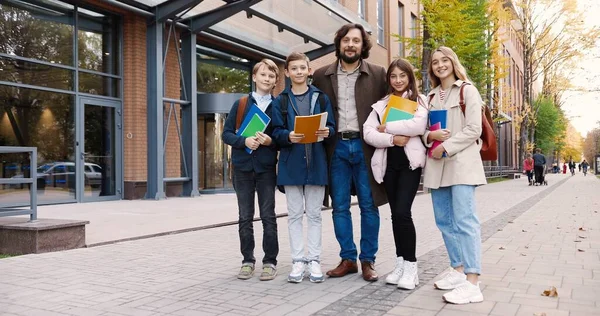 Χαρούμενα σχολιαρόπαιδα και μαθήτριες με πολύχρωμα copybooks που στέκονται με τον δάσκαλο στο δρόμο. Ευτυχισμένος καθηγητής που αγκαλιάζει τους μαθητές του. Καυκάσια κορίτσια και αγόρια μαθητές κοντά στο σχολείο — Φωτογραφία Αρχείου