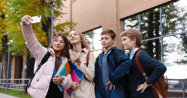 Retrato de niños felices de la escuela tomando fotos de selfies en el teléfono inteligente con amigos mientras están de pie al aire libre y haciendo caras divertidas. Compañeros de clase caucásicos tomando fotos en el teléfono celular y sonriendo —  Fotos de Stock