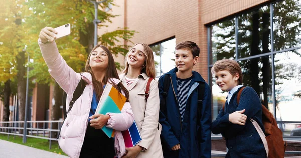 Portret van vrolijke schoolkinderen die selfie foto 's maken op smartphone met vrienden terwijl ze buiten staan en grappige gezichten trekken. Kaukasische klasgenoten nemen foto 's op mobiele telefoon en glimlachen — Stockfoto