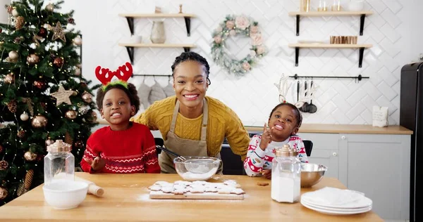 African American joyful mom with little pretty kids standing at table in cozy kitchen at home while making xmas cookies and biscuits. Happy family smiling to camera on Christmas Eve. New Year concept