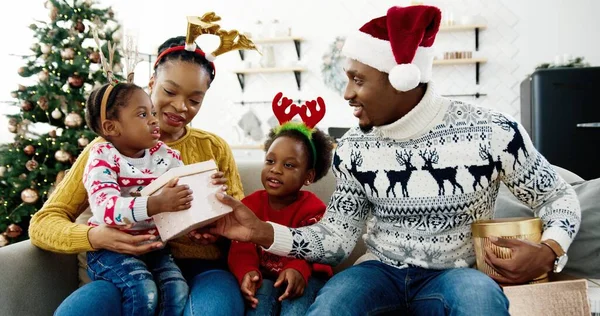 Portret van een gelukkige Afro-Amerikaanse vader in kerstmuts die kerstcadeaus geeft aan familie terwijl hij bij een versierde kerstboom thuis zit. Nieuwjaarsviering, kerstcadeaus. Vakantieconcept — Stockfoto