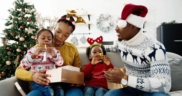Portret van nieuwsgierige Afro-Amerikaanse kinderen openen geschenkdozen terwijl ze samen zitten met gelukkige ouders in versierd huis in de buurt van mooie kerstboom oudejaarsavond. Vakantie viering xmas huidige concept — Stockfoto