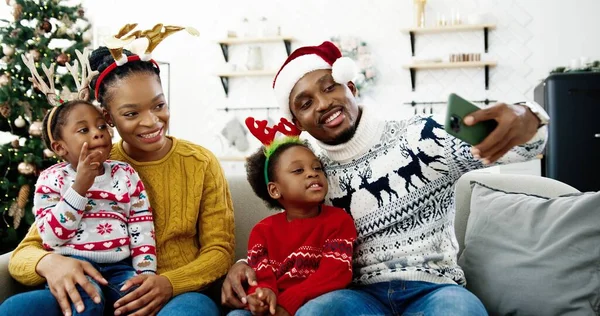 幸せなアフリカ系アメリカ人の家族の肖像写真をスマートフォンで撮影し、居心地の良いクリスマス装飾の家で笑顔。パパでサンタの帽子撮影セルフィー写真携帯電話で子供と妻 — ストック写真