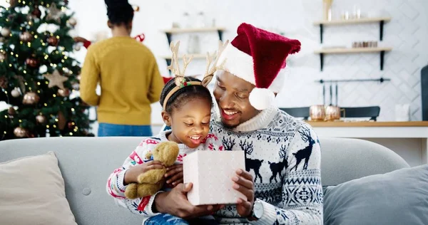 幸せなアフリカ系アメリカ人の小さな子供のクローズアップは、装飾された部屋でサンタの帽子のお父さんと座っている間にクリスマスプレゼントボックスを開きます。背景にクリスマスツリーを飾るママと子供。クリスマスプレゼントコンセプト — ストック写真