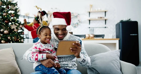 幸せなアフリカ系アメリカ人の小さな子供が、装飾された部屋でサンタの帽子に座って、タブレットで漫画を見ています。背景にクリスマスツリーを飾るママと子供。クリスマスのコンセプト — ストック写真