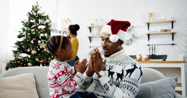 小さな子供に歌を教え、クリスマスハウスのソファに座っている間に遊んでいる父親の肖像画を閉じます。アフリカ系アメリカ人の母親と子供が背景に新年の木を飾る。クリスマスのコンセプト — ストック写真