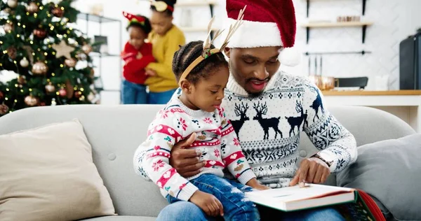 Close-up portret van vader lezen boek aan kleine vreugdevolle kind en spelen tijdens het zitten in gezellige moderne huis. Afro-Amerikaanse moeder en kind versieren nieuwjaarsboom op de achtergrond. Kerstconcept — Stockfoto