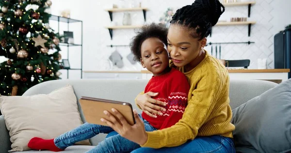 Afro-Amerikaanse vrolijke moeder en kind zitten op de bank in ingerichte moderne kamer in de buurt van kerstboom en kijken iets op tablet. Vrouw tikt op het apparaat en toont het aan het kind. December vakantie — Stockfoto