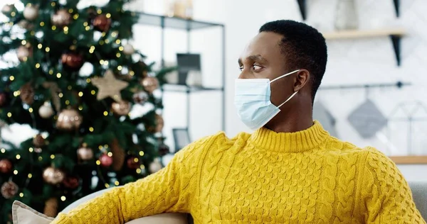 Primer plano retrato de guapo hombre afroamericano feliz en máscara médica sentado en el sofá en casa cerca del árbol de Navidad decorado con luces centelleantes y mirando a la cámara de buen humor. Concepto de Navidad —  Fotos de Stock