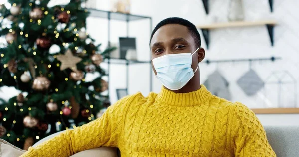 Primer plano retrato de guapo hombre afroamericano feliz en máscara médica sentado en el sofá en casa cerca del árbol de Navidad decorado con luces centelleantes y mirando a la cámara de buen humor. Concepto de Navidad —  Fotos de Stock