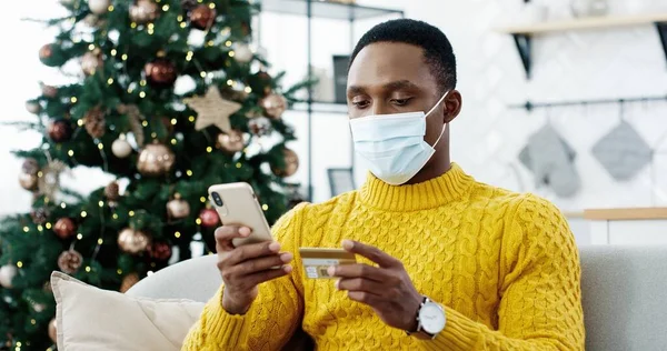 Primer plano retrato de hombre afroamericano feliz en la máscara sentado en la habitación cerca del árbol de Navidad con luces y la compra en línea en el teléfono inteligente que paga con tarjeta de crédito pidiendo regalos. Año Nuevo e-shopping —  Fotos de Stock