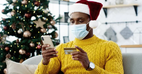 Hombre afroamericano con máscara médica y sombrero de santa cerca de árbol de Navidad decorado brillante celebración de tarjeta de crédito y teléfono inteligente haciendo transferencia de dinero en línea. Venta de Navidad, comprar regalos en internet, e-commerce —  Fotos de Stock