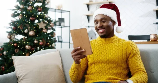 Portrét Afroameričana veselý mladý muž nosí Santa klobouk sledování videa on-line na tabletu a směje se uvnitř místnosti s pěknými vánoční zdobené stromeček. Koncept období nového roku — Stock fotografie