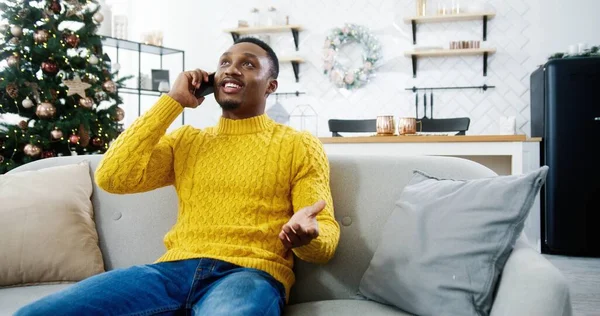 Excited African American happy man sit near decorated glowing tree enjoying nice talk with friend on Christmas Eve on smartphone. Male chatting on cellphone at home. New Year concept