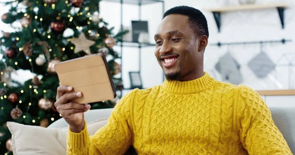 Närbild porträtt av afroamerikanska glad man talar på videosamtal på surfplatta med vän och har bra samtal skicka hälsningar sitter nära julgran i rummet. Begreppet nyår — Stockfoto