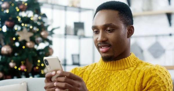 Primer plano retrato del alegre hombre afroamericano en suéter amarillo sentado en casa con el árbol de Navidad brillante y mensajes de texto en el teléfono inteligente enviar saludos y deseos a los amigos Feliz Año Nuevo concepto — Foto de Stock