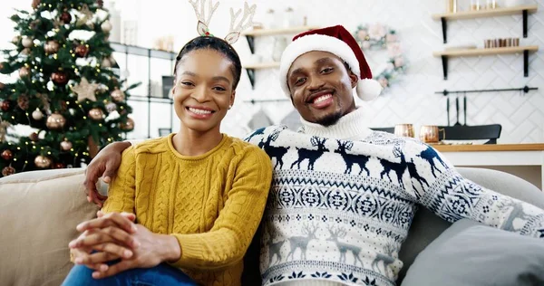 Portret van een gelukkig Afro-Amerikaans getrouwd stel, zittend in een kamer met versierde gloeiende kerstboom, kijkend naar de camera en glimlachend in een goede kerststemming. Gelukkig nieuwjaarsconcept. Gezinsvakantie — Stockfoto
