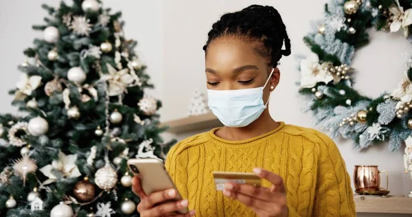 Primer plano retrato de mujer alegre en la máscara de hacer la compra de la venta de Navidad en línea en el teléfono inteligente con la cara feliz. Mujer afroamericana alegre comprando regalos en internet y pagando con tarjeta de crédito e-commerce —  Fotos de Stock