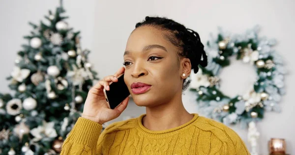 Κοντινό πορτρέτο της Αφροαμερικανής χαρούμενης νεαρής γυναίκας που μιλάει στο κινητό της σε διακοσμημένο δωμάτιο με χριστουγεννιάτικο στολισμό σε καλή διάθεση με φίλο και χαιρετισμό με διακοπές. Πνεύμα των Χριστουγέννων. Χαρούμενες διακοπές — Φωτογραφία Αρχείου