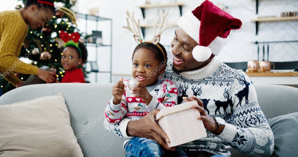 Close up of happy African American little kid opens xmas present box while sitting with dad in santa hat at decorated room. Mom and child decorating Christmas tree on background. Xmas gifts concept