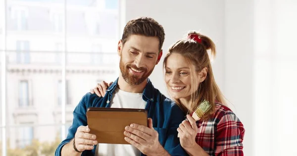 Close-up portret van vrolijke jonge getrouwde echtpaar man en vrouw staan in een nieuw appartement, tikken en surfen online op tablet, kiezen en bespreken van nieuw ontwerp van huis. Huisreparatie — Stockfoto