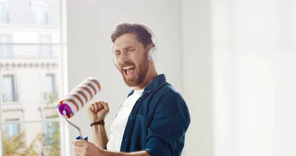 Närbild av glada kaukasiska rolig man lyssna sång i hörlurar, dans, flytta och sjunga till roller borste under hem reparation. Renovering och förbättring Man har roligt när man renoverar hus — Stockfoto