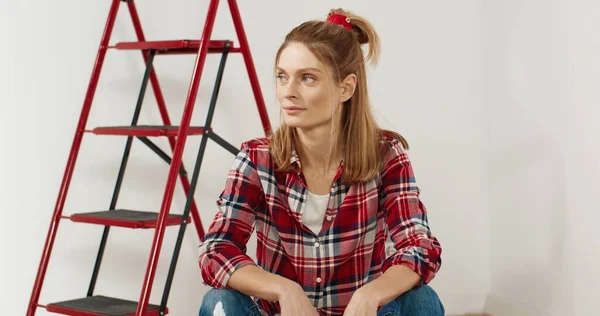 Retrato de joven joven alegre caucásica hermosa sentada en el apartamento cerca de la escalera, mirando a la cámara y sonriendo de buen humor con la cara feliz solo. Concepto de renovación y reparación del hogar — Foto de Stock