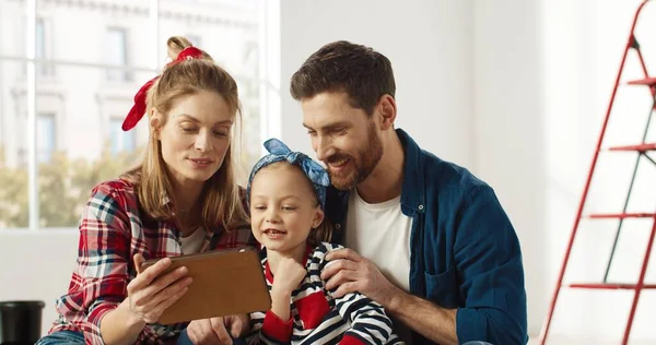 Gozosos padres caucásicos sentados con una linda niña preescolar charlando, tocando y navegando en línea en el dispositivo de la tableta, eligiendo una nueva decoración de la habitación en Internet juntos. concepto de reparación del hogar —  Fotos de Stock