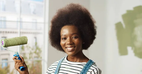 Retrato de sorrir jovem mulher afro-americana feliz olhando para câmera segurando pincel de rolo de pintura na mão em pé no quarto enquanto renovando e redecorando casa. Conceito de reparação e melhoria — Fotografia de Stock