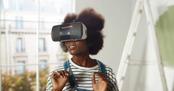 Zbliżenie Afroamerykanka młoda kobieta stoi w domu w okularach VR i przesuwanie z rąk w powietrzu za pomocą nowoczesnych innowacji naprawy i poprawy domu. Technologia rzeczywistości wirtualnej — Zdjęcie stockowe
