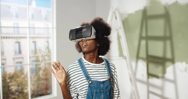 Close up da jovem afro-americana fica em casa vestindo óculos VR e deslizando rolagem com as mãos no ar usando inovações modernas reparar e melhorar a casa. Tecnologia de realidade virtual — Fotografia de Stock
