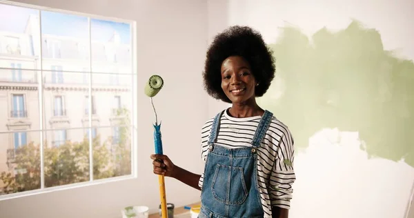 Retrato de sorrir jovem mulher afro-americana feliz olhando para câmera segurando pincel de rolo de pintura na mão em pé no quarto enquanto renovando e redecorando casa. Conceito de reparação e melhoria — Fotografia de Stock