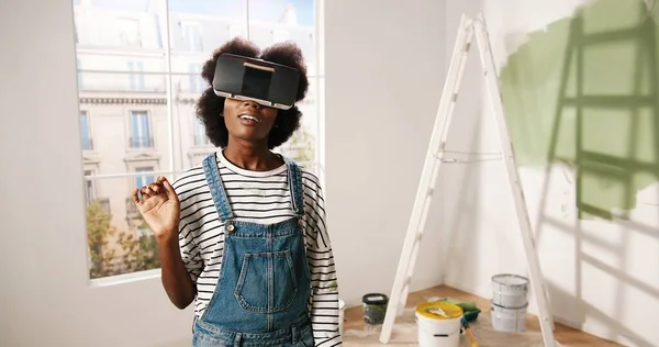 Retrato de afro-americano jovem impressionado mulher animado fica no quarto vestindo óculos VR e olhando para o novo apartamento renovado redesenhado usando inovações modernas e tecnologia de realidade virtual — Fotografia de Stock