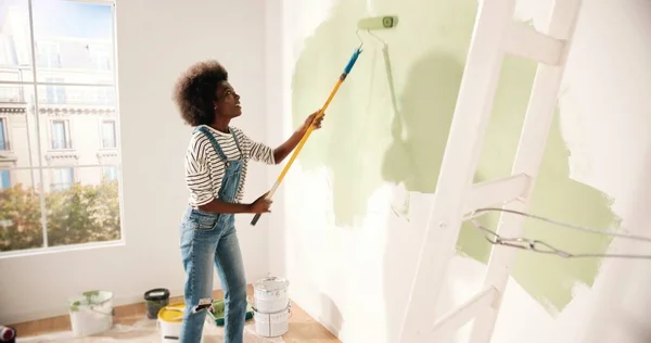 Ung afroamerikansk kvinna dansar och målar vägg med rullborste när du renoverar lägenhet. Bakre delen av honan har kul att möblera om hemma, renovera och förbättra Reparation och dekoration koncept — Stockfoto