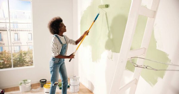 Young African American woman dancing and painting wall with roller brush while renovating apartment. Rear of female having fun redecorating home, renovating and improving Repair and decorating concept