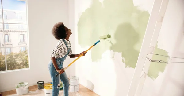 Jovem afro-americana dançando e pintando parede com escova de rolo ao renovar apartamento. Traseira da fêmea se divertindo redecorando casa, renovando e melhorando o conceito de reparação e decoração — Fotografia de Stock