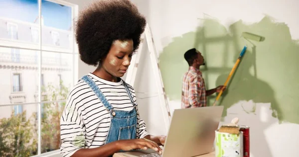 Retrato de mulher afro-americana bonita feliz digitando no laptop no quarto durante o reparo em casa falando com o marido que está pintando paredes em cor de azeitona com escova de rolo. Renovação e remodelação da casa — Fotografia de Stock