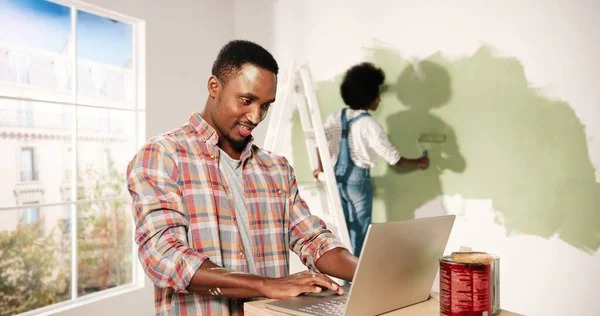 Retrato de homem afro-americano alegre digitando no laptop no quarto durante o reparo em casa falando com a esposa bonita que está pintando paredes em nova cor com escova de rolo. Renovação e remodelação da casa — Fotografia de Stock